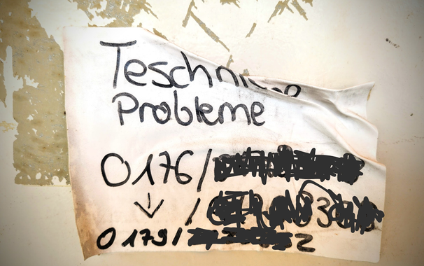 Ein Zettel an einem Münzautomaten bietet Hilfe "Teschniche Probleme" an (mit verbuchselten Wechstaben). 
Darunter verschiedene Handynummer, die teils mit Pfeilen aufeinander zeigen. Der Zettel klebt auch eher leicht zerknüllt an der Wand bzw. ist halb losgelöst heruntergerutscht. 

PS: Die Telefonnummern wurden natürlich auf dem Foto nachträglich unkenntlich gemacht...