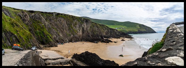 Panoramic photograph of Couminole Beach, County Kerry, Ireland, showing a secluded sandy cove bordered by dramatic, steep cliffs with patches of green grass, a few parked cars near the entrance, people scattered on the golden sand and in the surf, and a stone wall leading down to the beach under a partly cloudy sky.
