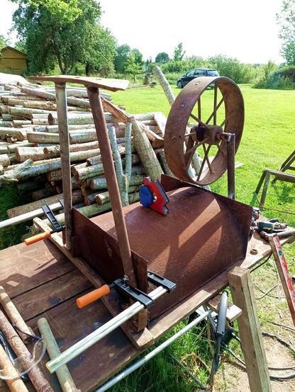 The wheelbarbie mid construction. The trough is upside down on a table, the spoke steel rimmed wheel already welded in place on two angled legs welded to an axle and the other leg has been manufactured from a plow share welded to two more pipes at an angle. This is currently held to the trough with two large C-clamps. Some welding magnets are idling on top of the trough and various tools are around. A stack of logs is behind and a parked car in front of a garden.