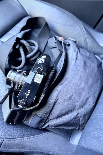 A photo of black 35mm rangefinder camera and a small sling bag sitting on a car seat.