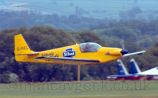 Slightly grainy side view of a bright yellow, single-engined light aircraft flying from left to right at a very low altitude, with it's fixed undercarriage mounted on thin stalks coming from the middle of the wings.
The plane is mostly a bright yellow, with blue "Utterly Butterly" titles on the rear fuselage, as well as a blue cross-like logo with white "St Ivel" text.
The blue registration "G-IVEL" is on the tail.
Grass and trees fill the background leading up to rolling hills covered in fields in the distance, with a white, blue and red military fighter jet in the bottom right corner.