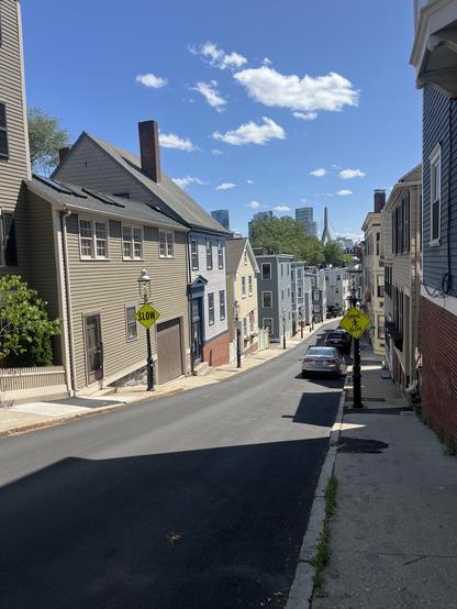 A row of various early 19th homes down a narrow descending street. There is a blue sky and a few small white clouds