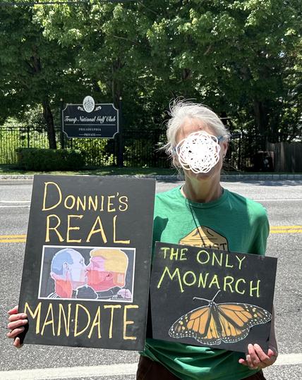 Sign in the background reads
Trump National Golf Course
Philadelphia
‐---‐-----------------
PRIVATE

Foreground 
Across the road, a protester holds a sign that reads 
Donnie's real Man-Date
On black poster board with the iconic image" Putin kissing Trump" graffiti from a cafe exterior wall which appeared in Latvia, 2016, and another sign on black poster board that reads THE ONLY MONARCH with an image of a butterfly.

Trump s*** hole is an hour from Philadelphia, in Pine Hill, NJ