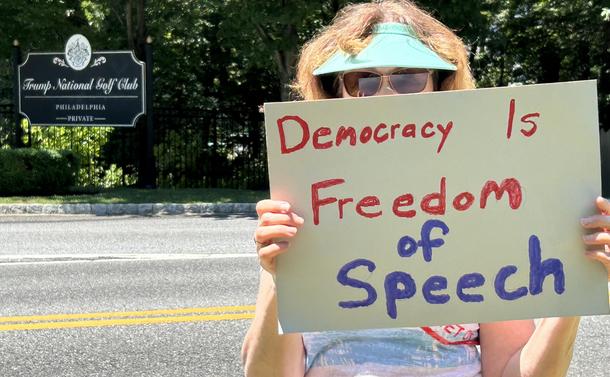 Sign in the background reads
Trump National Golf Course
Philadelphia
‐---‐-----------------
PRIVATE

Foreground 
Across the road, a protester holds a sign that reads 
DEMOCRACY IS
FREEDOM
OF
SPEECH