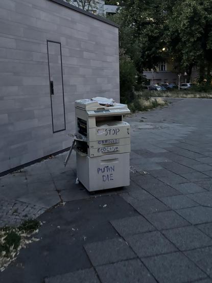 Büro-Kopierer vor einer öffentlichen Toilette stehend, auf einem Bürgersteig in Berlin Neukölln mit verschiedenen Parolen bemalt.