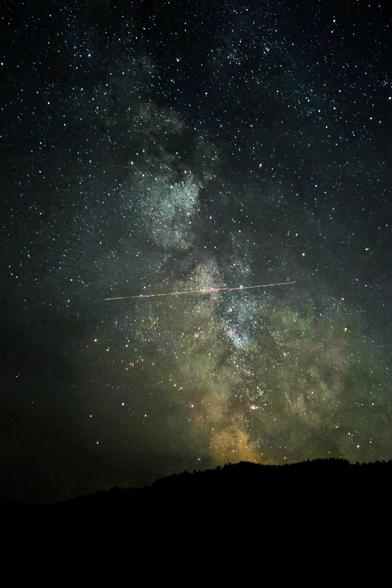 The Milky Way rises in a star-filled sky above the black shape of a forested hill. The galactic center, peering over the hill’s crest, glows a deeper red. An airplane’s light trail, caught in long exposure, appears to cross the galactic river perpendicularly from end to end.