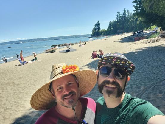 My husband and I posing in front of Third Beach. There's greenery and the ocean in the background along with folks lounging and the beach and preparing kayaks.