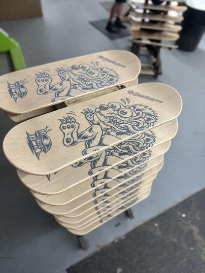 Stack of custom wooden skateboard decks (balance boards) featuring Honeybadger-themed artwork with an illustrated badger stomping on a bug design, displayed in a skateboard design workshop.