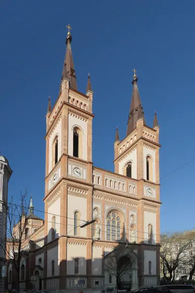 The image depicts a church building with a distinctive architectural style, featuring two tall towers with pointed roofs and decorative elements. The structure is characterized by its red and light-colored brickwork, large arched windows, and intricate details, suggesting a Gothic or Neo-Gothic influence. The clear blue sky in the background enhances the prominence of the building. If you need specific details about its history or location, feel free to ask!

Image Credits: Wikimedia / Thomas Ledl / CC BY-SA 3.0