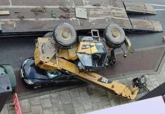 Photo of a CAT construction vehicle that has fallen off a truck bed onto a car parked on the side of the street.