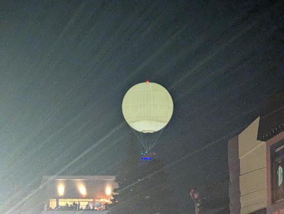 A glowing white orb atop a tower, against a night sky.  Buildings, one with a lit balcony, are visible along the bottom of the picture.