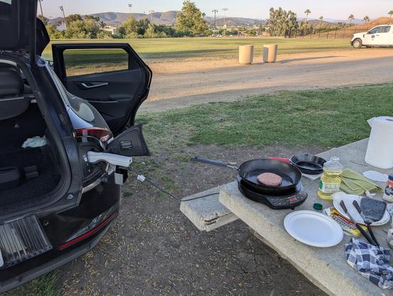 The back of a KIA EV6 with the hatch open. A V2L adapter is plugged into the charging port. An induction cooktop is sitting on top of a concrete picnic table, plugged into the adapter. On the cooktop, a carbon steel skillet cooks a burger patty