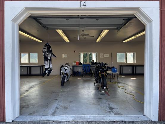Picture looking into a garage bay from outside. There are two motorcycles side by side. There’s a motorcycle suit on a hanger to the left. There’s a workbench and window in the back.