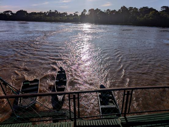 Die Aufnahme zeigt einen Fluß mit tropischer Vegetation am Ufer. Die tiefstehende Sonne, die nicht im Bild ist, wird im Wasser reflektiert. 
Der Betrachter steht am Heck eines Schiffs und blick hinab auf drei Beiboote, die es hinter sich her zieht.