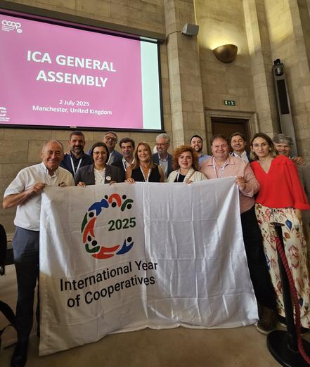 Un grup de persones cooperativistes sostenen una pancarta amb el logotip i la inscripció de l'Any Internacional de les Cooperatives, 2025. De fons, una pantalla indica que es troben a l'Assemblea General de l'Aliança Cooperativa Internacional (ICA, per les sigles en anglès).