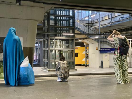 Photo taken at the station Ostkreuz: Three people, probably women, are waiting for the train. They do not Senn to know each other but they are all wearing different kinds of overalls so their bodies are covered in one kind of cloth.