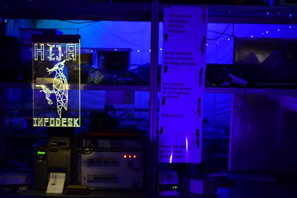 Links ein leuchtendes Schild "HOA Infodesk", daneben fünf Schilder übereinander "Bitte keine sinnlosen Schilder drucken", "Bitte wirklich keine sinnlosen Schilder drucken", "Das Drucken sinnloser Schilder ist strengstens untersagt", ""Echt jetzt?", "Immerhin haben wir es nicht lammentiert"