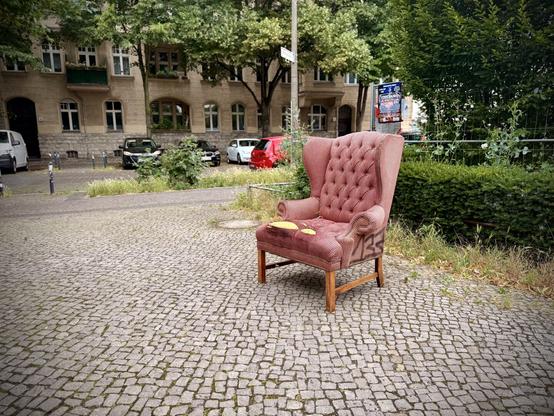 Ein alter roter Polstersessel steht auf Kopfsteinpflaster, umgeben von Grünpflanzen und geparkten Autos
