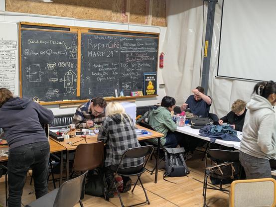 A group of people in the hacker space. There's a blackboard and tables of activities