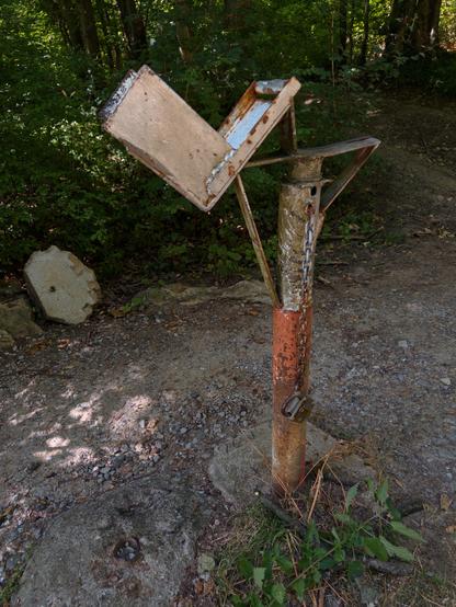 Eine verlassene Metallstruktur, die einem verwitterten Briefkasten ähnelt, der auf einer rostigen Stange montiert ist, umgeben von Grün und unebenem Boden. Eine Kette verriegelt die Stange am Boden, was darauf hindeutet, dass sie nicht mehr verwendet wird.