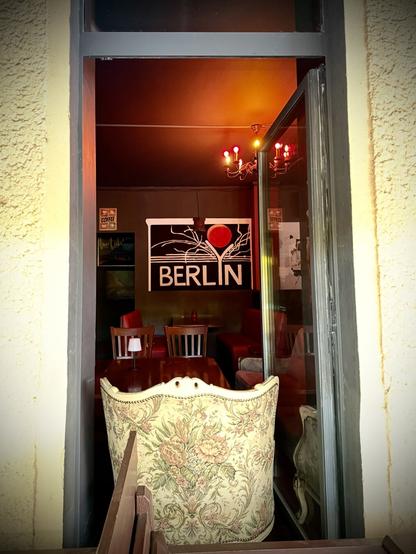 Blick in eine Bar durch ein bodenlanges Fenster, blockiert von einem gemusterten, alten Sessel, im Hintergrund hängt ein Schild auf dem BERLIN steht