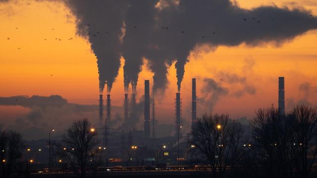 Image shows smoky industrial chimneys above a city skyline. The sun is setting in the sky highlighting a stratum of smog.