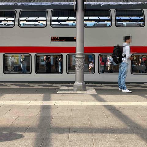 Bahnhof. Zugausschnitt. 2 stöckig. Links steht ein Mann. In der Mitte ein silbergrauer Pfeiler (alt u. restauriert) mit Ornament. Der Zug ist hellsilbergrau mit rotem Streifen über den Fenstern des ebenerdigen Geschosses. Es gibt weiße Markierungen auf dem im Vordergrund liegenden Bahnsteigs längs zur Bahnsteinkante.