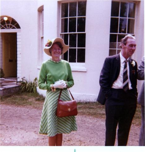 Outside a white building, the main subject of the photo is a 48-year-old woman, Mary. She's wearing a green dress that's solid green on the top half and green-and-white chevrons on the bottom half. She's also wearing a large, cream coloured hat, white gloves, and is carrying a brown handbag. A suited man is stood nearby.