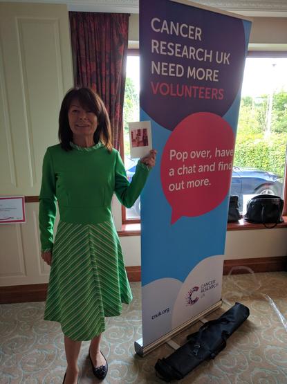 A woman of similar age to Mary wears the same green dress from the previous photograph, smiling for the camera whilst holding a copy of that photo. She's standing in front of a volunteer recruitment banner for Cancer Research UK.