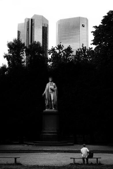 With the German Bank behind him, Friedrich Schiller's statue gives life advice to a young man of questionable social status sitting on a bench in front of him in the Taunusanlage, Frankfurt's green lung and “formerly” the site of an open drug scene near the main train station.

Just as the picture thrives on visual contrasts, the rear elevated level (Deutsche Bank) also represents a strong contrast in terms of content to the front lower level (young man of questionable social status at a problematic focal point). The middle level (statue of Friedrich Schiller) mediates, both visually and in terms of content.

Does the dark wall of the trees have a protective effect? If so, who does it protect and in which direction should it protect? Or does it hinder and make things impossible? In any case, Deutsche Bank, Schiller and the young man are connected characterized by similar tonal values. Well, and Schiller is on the side of the young man.