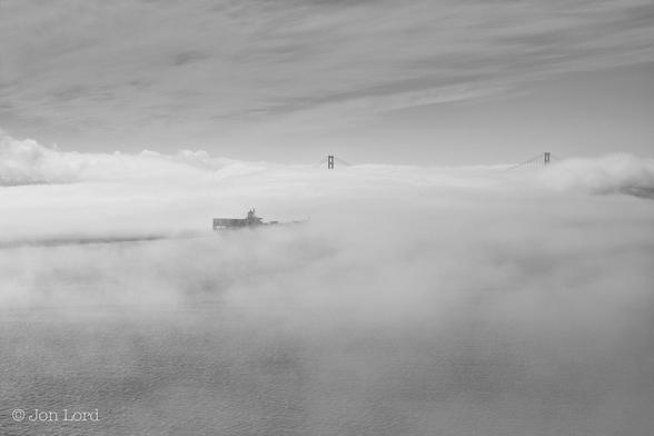 This is a black and white seascape photo of a cargo ship underway through a large bank of sea fog. Rising up two-thirds of the image is a bank of sea fog, broken in places so that the water below is visible. Slightly to the left of centre is a cargo ship underway from left to right. The aft superstructure is visible as is a small portion of the shipping containers stacked on the deck. Behind the ship is a dark trough marking the ship's passage through the sea fog. Ahead of the ship are the tops of the two towers of a large suspension bridge, some of the cables are visible as they descend into the mist. Above is a thin layer of high cirrus cloud.
The Location: The San Francisco Bay, the bridge: The Golden Gate (2017).