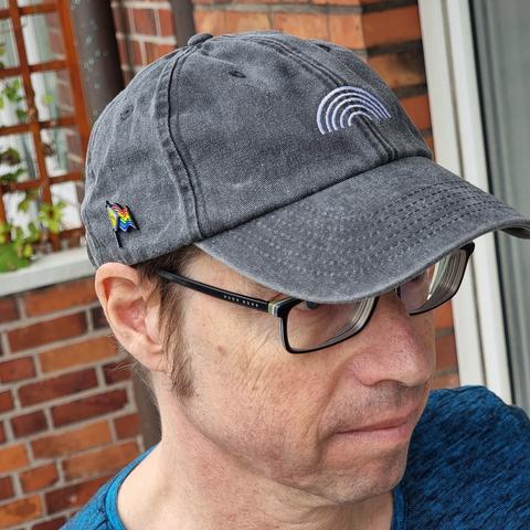 Mensch mit dunkler Brille, mittelblonden Koteletten und leichtem Bartschatten trägt Kappe aus grauem Baumwollstoff in einem ausgeblichenen unregelmäßigen Finish, wodurch die Kanten und die Konturen der Nähte besonders betont werden. Das Foto ist von schräg links oben, der freundliche Blick des Menschen geht nach rechts und leicht unten. Sein Schwimmbad-blau meliertes T-Shirt ist zu erkennen. Vorn auf der Kappe ist ein grafischer Regenbogen aufgestickt. Er besteht einfach nur aus bogenförmig übereinander gestapelten gewölbten Linien, die allesamt weiß statt farbig sind. An der uns halb zugewandten Seite der Kappe prangt ein kleiner farbiger Pin in Form einer stilisierten wehenden new Progress-Pride-Flagge. Im Hintergrund befinden sich rote Backsteine eines Balkons und einer Hauswand. Auch ein rötlich braunes Rankgitter umgeben von ein paar hellgrünen kleineren Blättern ist dort verschwommen im Bild. Ganz rechts durchschneidet der weiße Rahmen einer Fensterfläche im Hintergrund die Gesamthöhe des Bildes.