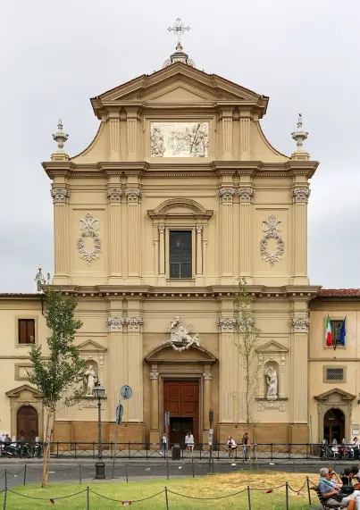 The image depicts a large, ornate building with architectural features characteristic of a church or religious structure. The façade is decorated with sculptures and reliefs, and there appears to be a central door flanked by a couple of windows. People can be seen sitting in front of the building, and there are trees and a fence in the foreground. Additionally, flags can be spotted, indicating a possible location in Italy. The overall atmosphere appears to be calm and serene, set against a cloudy sky.

Image Credits: Wikimedia / Sailko / CC BY 3.0