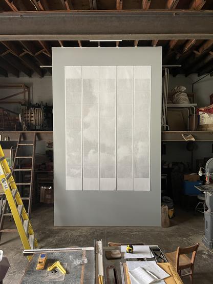 50 sheets of wide format computer paper hang in strips of 10 on the gray wall of a workshop. A faint, dithered image of clouds is visible on the paper. In the foreground is a table saw with papers and a book on its surface. Two ladders are visible on the left and the edge of a bandsaw can be seen on the right.