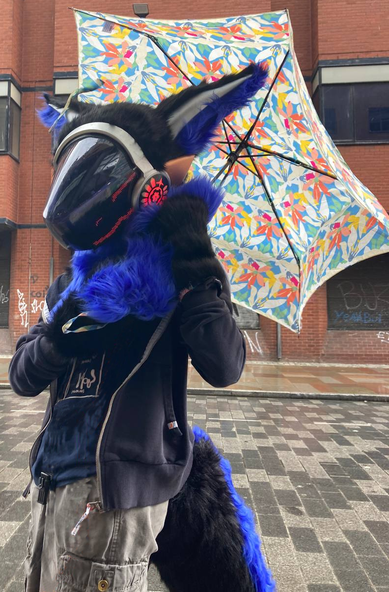 A photograph of a protogen partial fursuit with blue and black fur, bronze and silver metallic paints on plastics, red LEDs and a long bushy tail. It is a rainy day in a urban area and the fursuiter is holding an umbrella and facing to the left.
