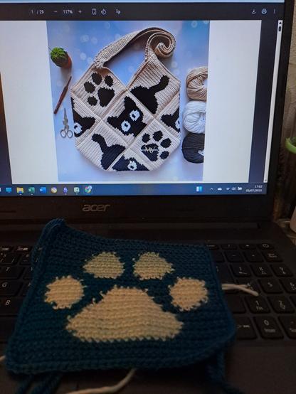 Le même carré au crochet posé sur un clavier d'ordinateur.  En fond, un modèle de sac au crochet composé de carrés sur le thème des chats.