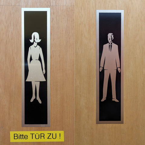 Die Abbildung zeigt zwei Türeingänge, die jeweils mit einem Hinweisschild versehen sind. Auf der linken Seite ist ein Schild mit einem stilisierten weiblichen Silhouette abgebildet, die ein Kleid trägt. Auf der rechten Seite ist ein Schild mit einem stilisierten männlichen Silhouette abgebildet, der einen Anzug trägt. Beide Schilder sind schwarz mit einem silbernen Rand und befinden sich auf einer Holztür. Unterhalb der beiden Schilder ist ein gelbes Schild mit der Aufschrift "Bitte TÜR ZU!" in schwarzer Schrift.