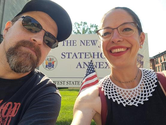 A middle-aged man on the left, with a beard and mustache, is wearing a cap and sunglasses.  To the right is a middle-aged woman whose dark hair is tied back in a bun.  She is wearing a black, sleeveless tee with a handmade white crocheted collar.  Her appearance resembles the late Supreme Court Justice, Ruth Bader Ginsberg.  A sign behind them reads, "The New Jersey Statehouse Annex."