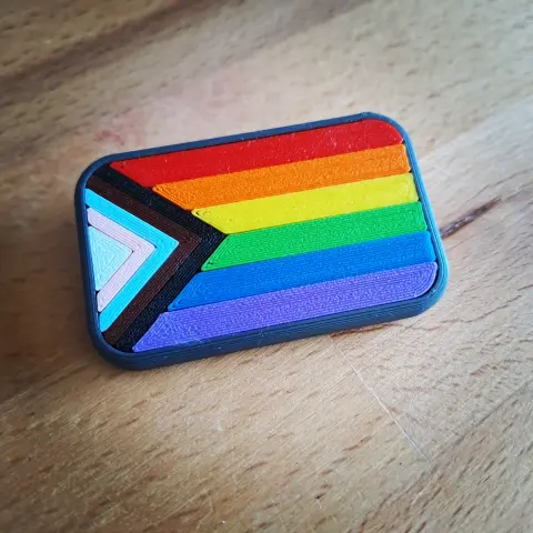 Face avant du badge représentant le drapeau arc-en-ciel incluant les couleurs de la transidentité et des personnes racisées.