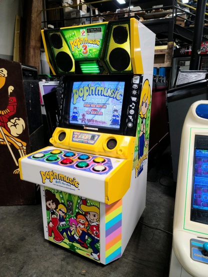 a pop'n music arcade cabinet in an auction house. it's an old but charming looking yellow upright cab design with a top section that has two speakers with yellow backlighting, a 'popn music 3' marquee, and cute little neon bars underneath it. it has a crt monitor with (unlit) lights with a mask on them to have little icons. the controller consists of nine large colorful buttons. it has character art on the sides and on the operator door in front