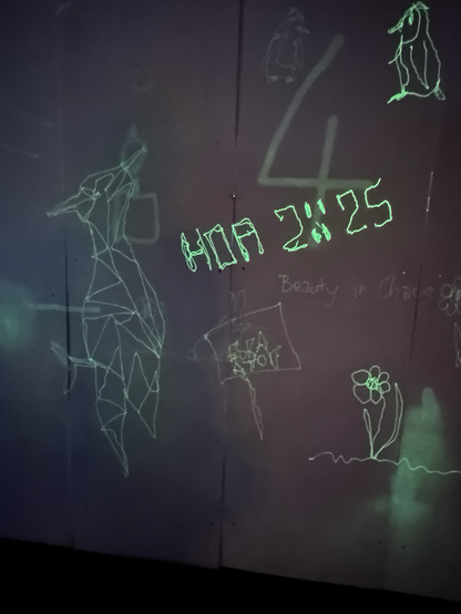 Weiße Wand mit uv-aktiven Farbauftrag, darauf mit UV-Taschenlampen gemalte Sachen, u.a. der Schriftzug HOA 2025 und der Specht vom Logo und eine Blume