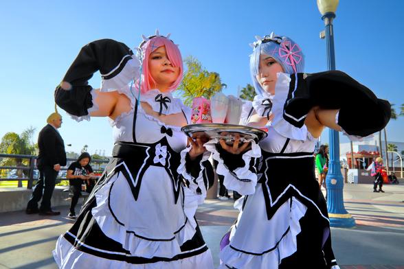 Two people cosplaying as maid characters.  Left is wearing a pink wig with left eye covered and right is wearing a blue wig with right eye covered.  Both are holding silver trays with beverages and shakes on them.  Their arms are up in a symmetrical pattern. Arms are up with sleeve covered hands near the face and armpits exposed as they are wearing tank top style white maid tops.
