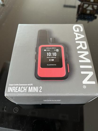 An image of a Garmin inReach Mini 2 satellite communicator packaging. The device is prominently displayed, featuring a red and black design with a screen showing the time and connection status. The packaging includes the brand name "GARMIN" and product description.