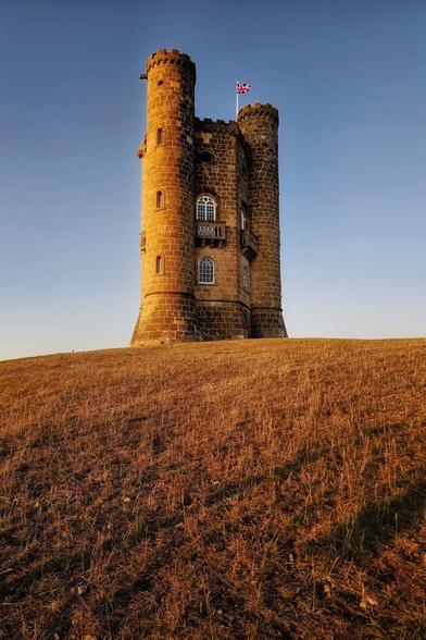 A low-angle shot of Broadway Tower, a tall, historic stone folly, standing prominently on a golden-brown grassy hill against a clear, pale blue sky at sunset. The warm, low sunlight illuminates the tower's stonework and the dry grass in the foreground, with a small British flag visible at the very top of the tower.