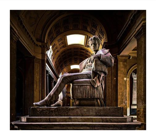 The photo is a statue of a young man seated on a chair, situated within a grand, classical-style hallway. The statue appears to be made of stone, and is a man in period clothing, possibly from the 18th century with a thoughtful or pensive expression. The hallway features arched ceilings with ornate detailing. The lighting is dramatic, highlighting the statue against the darker background of the hallway.