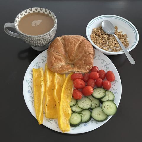A plate full of sliced cucumbers and sweet pointed peppers with grape tomatoes, a bowl of yogurt topped with some granola, and a croissant with almond coffee