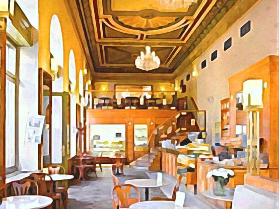 Innenansicht eines Cafés, im Vordergrund Tische, im Mittelgrund rechts eine Bar, im Hintergrund eine Art Zwischengeschoss in dem sehr hohen Raum, dass durch eine Treppe  erschlossen ist, auf diesem "Podest" stehen auch Tische. Die linke Seite des Raumes ist durch 5 hohe Bogenfenster belichtet, die rechte ist eine Wand mit div. Schränken