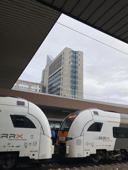 Ein Regionalzug. Zu sehen sind Anfang und Ende zweier aneinander gekoppelter Zugteile. Sie sind weiß. Durch das Dach des Bahnhofs  ist ein Hochhaus zu sehen.