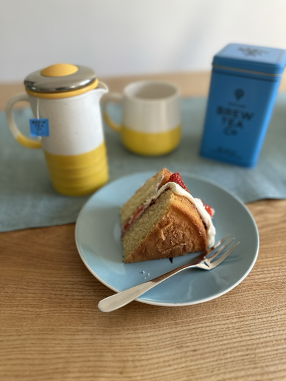 A picture of a slice of Victoria Sponge on a plate with a yellow and white teapot and mug set and a blue tea caddy