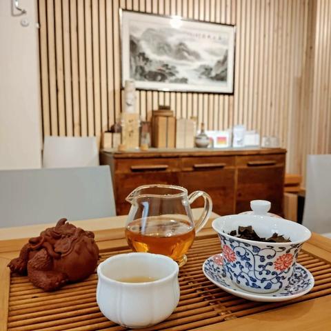 A gong fu cha water tray with a toad clay tea pet as well as a gaiwan, pitcher and cup. The tea in the pitcher is dark orange. In the background is an ink painting of Chinese mountains.